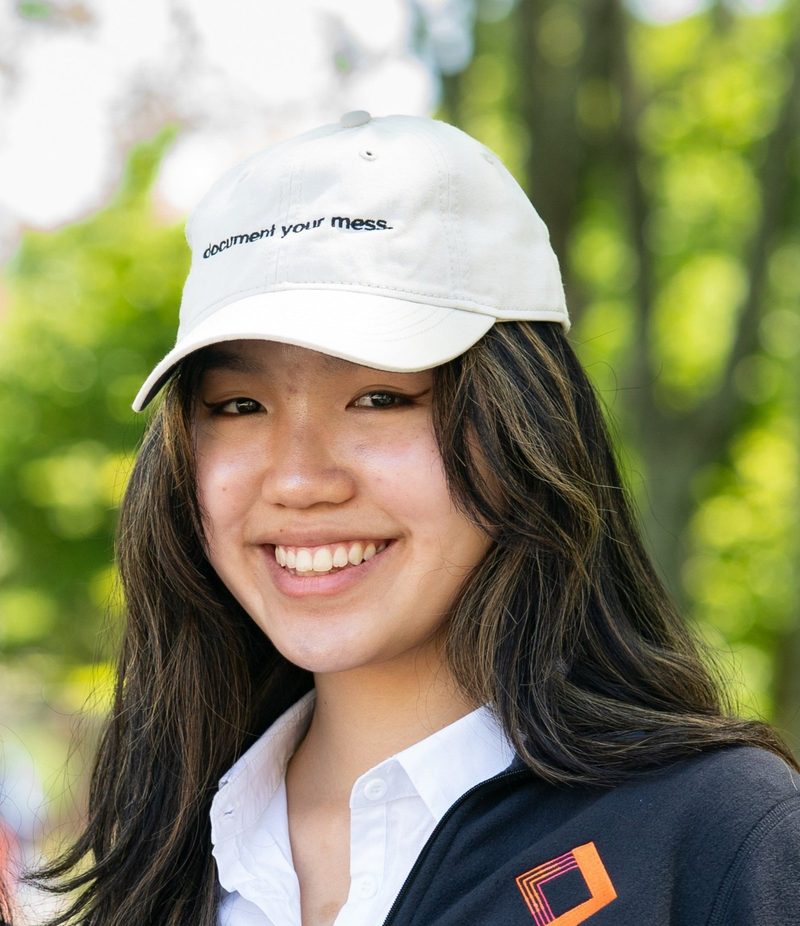 Chloe in a white cap that reads document your mess, under the trees